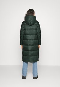 Kvinde iført en lang mørkegrøn pufferjakke med hætte, lyseblå jeans og sorte støvler, stående med ansigtet vendt mod en hvid væg.