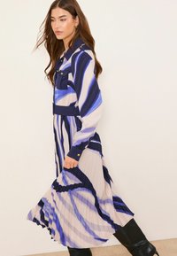 Langärmeliges, knöpfbares Kleid mit plissiertem Design. Farben: Marineblau und Creme mit abstrakten blauen Streifen. Schwarze kniehohe Stiefel.