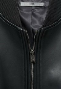 Chaqueta bomber de cuero negra con cuello acanalado, cuenta con un cierre metálico en la parte delantera y un forro interior suave. Etiqueta de la marca visible.