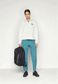 Naine seisab, samuti musta seljakoti käes, seljas valge flanellist sviiter, türkiissinised püksid ja mustad tossud, lihtsa heleda tausta ees.