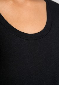 Juodas marškinėliai su plačia apvalia iškirpte, minkšta tekstūra ir lengvu audiniu, rodančiu lengvus audinio storio skirtumus.