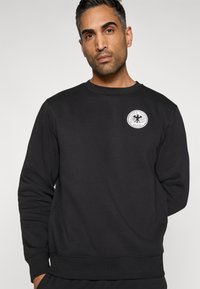 Melns džemperis ar apaļu kakla izgriezumu, ribotām aprocēm un apakšu, uz krūtīm ar apaļu baltu logotipu. Mīksts, biezs audums ar gludu tekstūru.