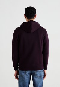 Felpa viola scuro con cappuccio regolabile, polsini e orlo a coste, abbinata a jeans blu chiaro. Tessuto con una texture liscia.