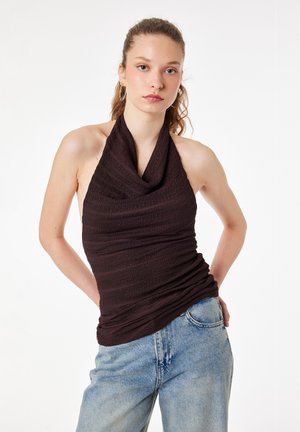 Jeune femme aux cheveux bouclés porte un top noir texturé sans manches à col halter et un jean bleu clair, debout les mains derrière le dos.