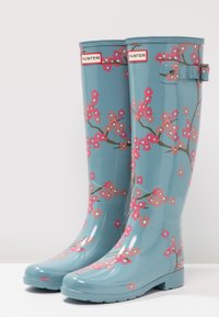 Botas de lluvia de goma azules con un acabado brillante, que cuentan con un diseño floral rosa y ramas marrones. Hay una correa ajustable en el lado.
