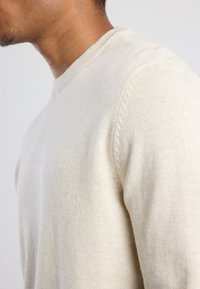 Maglione beige chiaro con un design a maglia testurizzata, caratterizzato da un collo rotondo e dettagli a costine sul collo e sulle cuciture delle spalle.