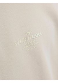 Un tejido de color beige claro y textura suave presenta un texto en relieve en blanco: "STUDIO COLL. 1990 JACK & JONES DESDE MIL NOVECIENTOS NOVENTA Colección."