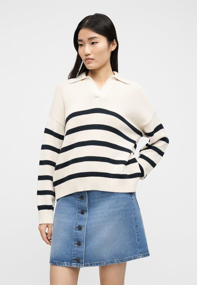 Γυναίκα που φοράει κρεμ πουλόβερ με μαύρες οριζόντιες ρίγες και γιακά, συνδυασμένο με μπλε denim φούστα με κουμπιά μπροστά, στέκεται μπροστά σε λευκό φόντο.