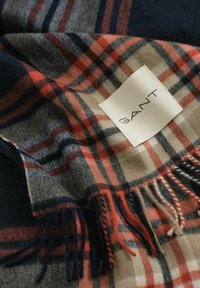 Geruite wollen sjaal in marineblauw, rood en beige tinten met franjes; heeft een GANT-logo label op een gladde stofoppervlak.