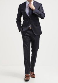 Costume bleu marine avec un blazer à un bouton et un pantalon à taille plate. Chemise bleu clair et cravate à motifs. Chaussures habillées marron. Design simple et ajusté.