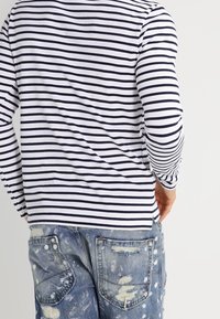 Langsleeve shirt met marineblauwe en witte horizontale strepen, gecombineerd met lichtblauwe versleten jeans met meerdere patches en rafelende randen.