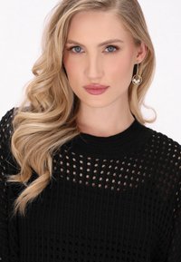 Zwarte gebreide trui met een losse, open geweven ontwerp. Het model heeft lang, golvend blond haar en draagt gouden oorringen met een parelaccent.