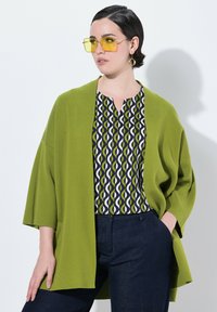 Zelený pletený cardigan přes vzorovanou blůzu s kruhovými vzory v zelené a námořnické modři, doplněný tmavě modrými džíny a žlutými slunečními brýlemi.