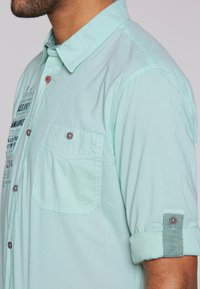 Camicia verde chiaro a maniche corte in cotone con tasca, dettagli in bottoni e polsini arrotolati con tessuto a contrasto.