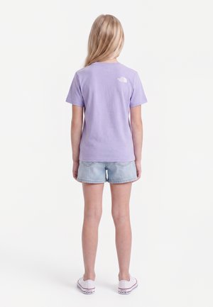 Barn med langt blondt hår iført en lyse lilla kortærmet top, lyse denimbukser og hvide sneakers, stående med ryggen til.