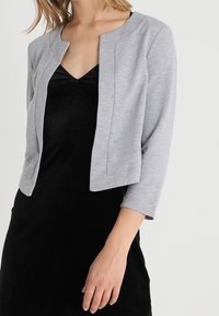 Femme portant une robe noire à décolleté en V avec une veste cropped ouverte sans col, de couleur gris clair, avec des manches trois-quarts.