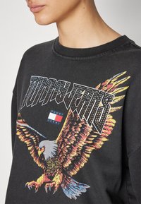 Zwarte sweatshirt met een groot adelaarsgrafiek in levendige kleuren, het logo "TOMMY JEANS" in vette letters en een klein merkembleem.