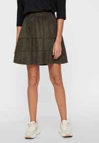 Olivově zelená, vícevrstvá sukně s texturovanou tkaninou a elastickým pasem, kombinovaná se světlými sportovními botami. Sofistikovaný a ležérní design.