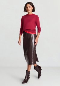 Roter Strickpullover über einem roten Oberteil, kombiniert mit einem glänzenden braunen Leder-Midirock und braunen Stiefeletten mit spitzen Zehen.