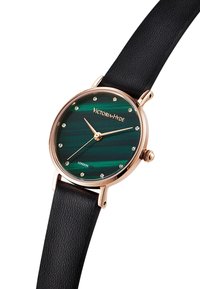 Okrągły zegarek z zieloną paskowaną tarczą, różowozłotymi akcentami i czarnym skórzanym paskiem. Wyposażony w znaki w postaci gemów oraz proste wskazówki godzinową i minutową.