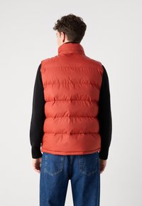Rød puffer vest med høy krage, quiltet design og en glatt tekstur, sammen med en svart langermet skjorte og blå jeans.