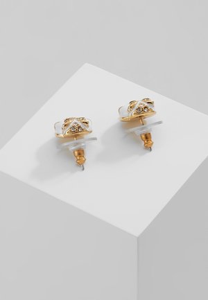 Paire de petites boucles d'oreilles clous dorées et blanches avec accents en cristal sur un bloc blanc sur fond gris.