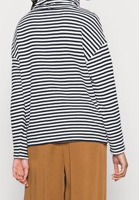 Pruhovaný top s dlouhým rukávem v námořnické modré a bílé, s volným střihem a vysokým límcem. Nošený s hnědými širokými kalhotami. Texturovaná látka.