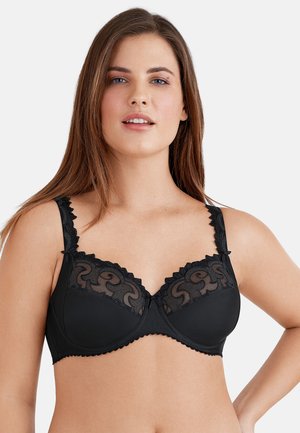 Frau trägt einen schwarzen Spitzen-Büstenhalter mit Bügeln, gezackten Trägern und bestickten Cups, steht vor einem einfarbigen hellen Hintergrund.