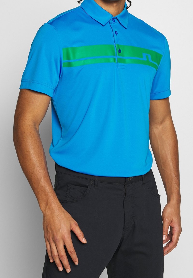 Polokošile s krátkým rukávem v jasně modré látce s hladkou texturou; má zelené horizontální pruhy přes hruď a límec na knoflíky.