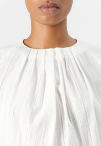 Hvit bluse med vertikale striper, strukturert stoff, høy hals, og plissédetaljer. Frontlukking er subtil med samlet stoff.