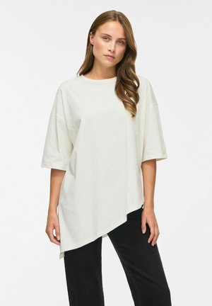 Ung kvinde med langt brunt hår iført en løs hvid asymmetrisk t-shirt og sorte bukser står foran en ensfarvet hvid baggrund.