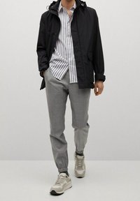 Zwarte lichtgewicht jas met capuchon, wit en grijs gestreept shirt, grijze tapered broek en beige sneakers. Kenmerken zijn onder andere een rits aan de voorkant en zakken.