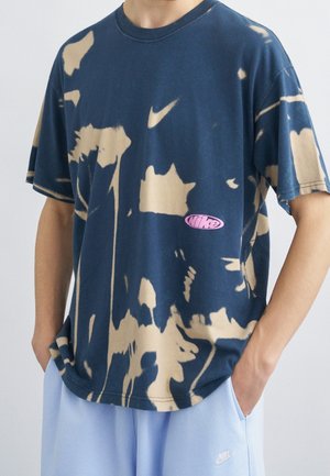 Personne portant un t-shirt Nike tie-dye bleu et beige avec un logo rose, associé à un short Nike bleu clair, mains dans les poches.