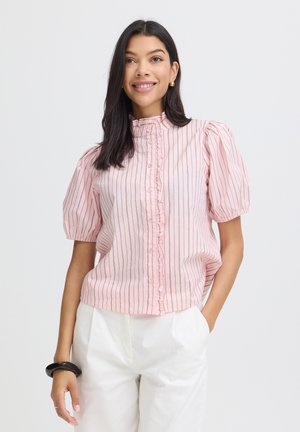 Žena s tmavými vlasmi, oblečená v ružovej pruhovanej blúzke s nadýchanými rukávmi a bielych nohaviciach, usmievajúca sa s jednou rukou v vrecku.