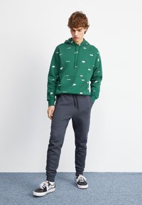 Grüne Kapuzenjacke mit weißen Logo-Mustern, Kängurutasche, kombiniert mit dunkelgrauen Jogginghosen. Die Schuhe bestehen aus schwarz-weißen karierten Vans.