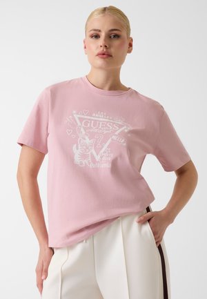 Femme aux cheveux blonds portant un t-shirt rose Guess à motif blanc et un pantalon crème avec une bande foncée, debout devant un fond uni.