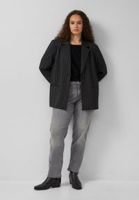 Blazer noir à rayures avec épaules rembourrées, grandes poches et manches à longueur de coude. Associé à un jean gris et des bottines noires à talons.