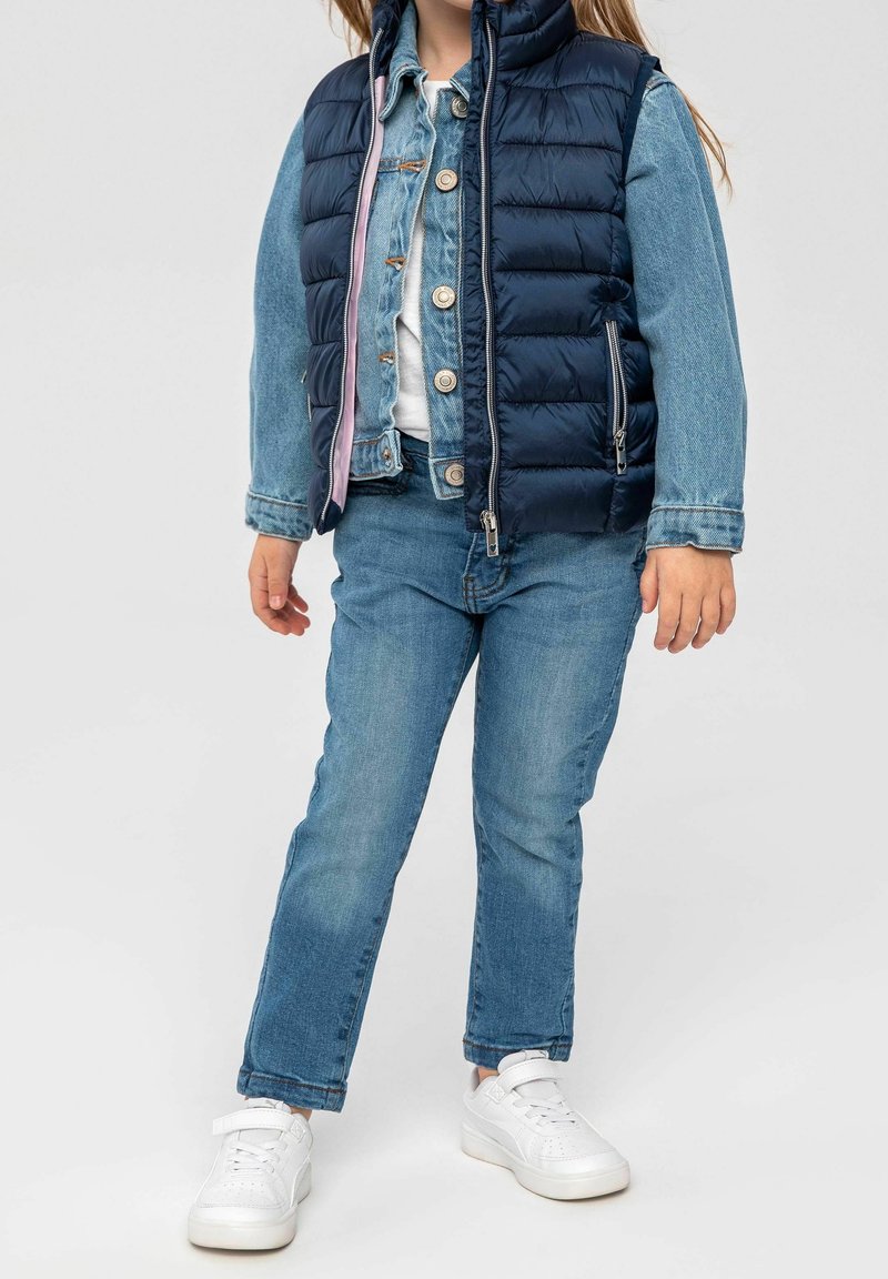 Gepolsterte marineblaue Weste über einer hellen Denimjacke mit Knopfverschluss, kombiniert mit hellblauen Jeans und weißen Sneakers, um einen Lagenlook zu kreieren.