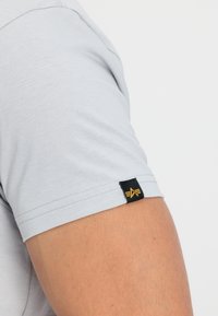 Svetlo siva majica s kratkimi rokavi iz bombaža, ki ima črno oznako z zlatimi detajli na levem rokavu. Gladka tekstura, klasičen kroj.