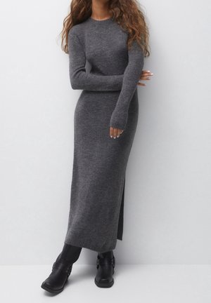 Femme portant une robe en maille grise à manches longues avec une fente latérale, associée à des bottines noires, debout devant un fond blanc uni.