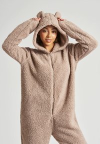 Brun, blød fleece onesie med hætte, der har bjørneører. Designet inkluderer en frontlynlås og lange ærmer, hvilket fremhæver den bløde tekstur.
