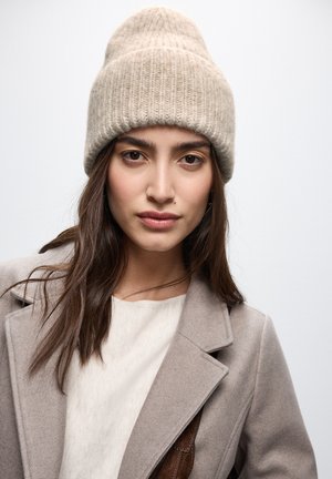 Beige strikket hue med ribbet tekstur, der har en foldet skygge, båret sammen med en lysfarvet top og en grå frakke, mod en ensfarvet baggrund.