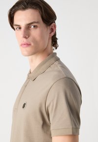 Beige polo shirt made of smooth fabric, featuring a ribbed collar and a subtle logo patch on the left chest. Short sleeves with fitted cuffs.