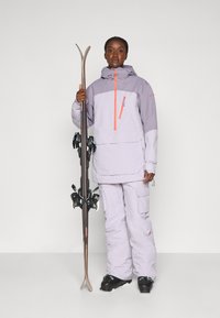 Lyžařský outfit ve světle fialové a šedé barvě s oranžovým zipem, obsahující bundu s kapucí a cargo kalhoty. Osoba drží lyže a spojky.