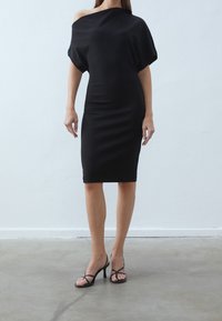 Zwarte off-shoulder jurk van gladde stof, nauwsluitend model, tot op de knie, gecombineerd met zwarte sandalen met hoge hak en bandjes.