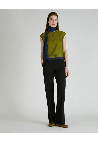 Maglietta verde a maniche corte con motivo geometrico, abbinata a pantaloni neri. La maglietta presenta disegni geometrici e un accento con sciarpa blu.
