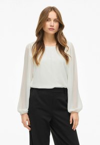 Blusa bianca trasparente con maniche lunghe e arricciate; scollatura rotonda. Abbinata a pantaloni neri a vita alta per un look a contrasto.