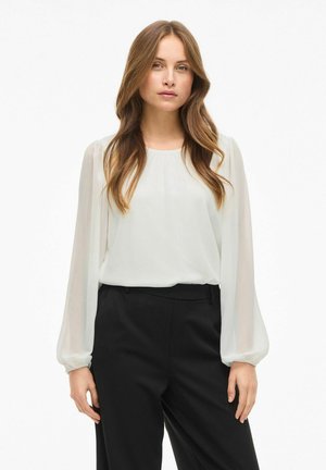 Blusa bianca trasparente con maniche lunghe e arricciate; scollatura rotonda. Abbinata a pantaloni neri a vita alta per un look a contrasto.