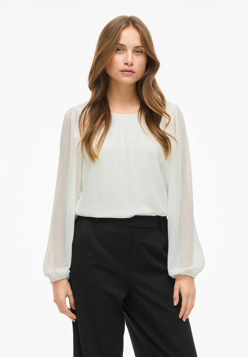 Blusa bianca trasparente con maniche lunghe e arricciate; scollatura rotonda. Abbinata a pantaloni neri a vita alta per un look a contrasto.
