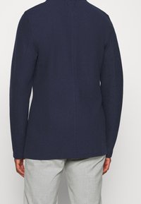 Navy blue jacket with a textured fabric, long sleeves, and a straight cut. Features a seam down the center and sits at the hip.